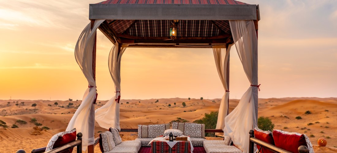 Arabisches Wüstencamp bei Sonnenuntergang mit einem offenen Zeltpavillon in traditionellem Design. Im Inneren liegen orientalische Teppiche und gemütliche Sitzpolster rund um einen niedrigen Tisch. Leichte Vorhänge wehen im Wind, im Hintergrund erstreckt 