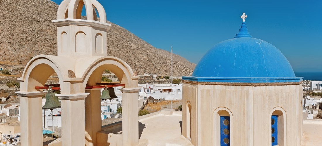 Kirche in Emporio, Santorini: freistehender Glockenturm mit drei Glocken neben Kapelle mit blauer Kuppel vor den Berghängen
