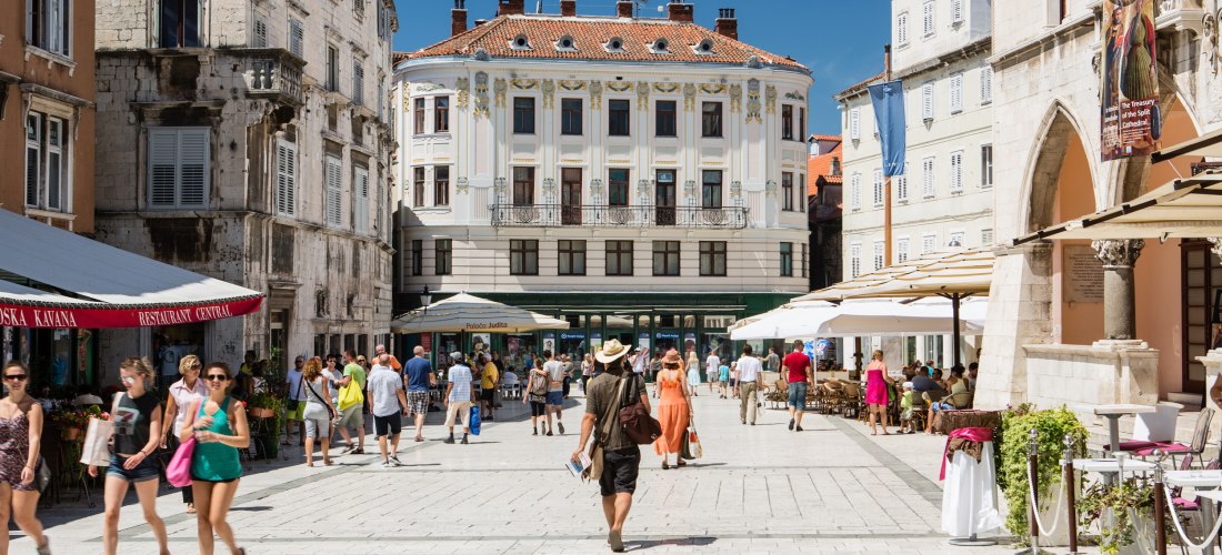 Kroatien - Split elebter Platz in der Altstadt von Split mit historischen Gebäuden, Straßencafés und vielen flanierenden Menschen bei Sonnenschein. Im Hintergrund ein prachtvolles Gebäude mit verzierten Fensterrahmen.