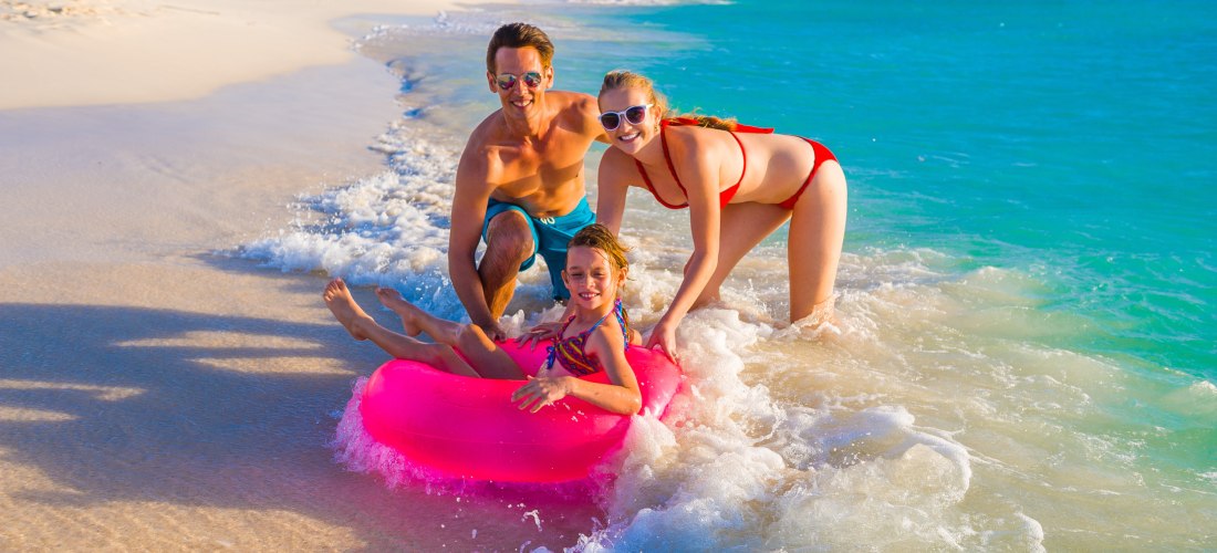 Eine lachende Familie am Strand von Aruba: Ein Vater und eine Mutter spielen mit ihrer Tochter, die in einem pinken Schwimmring in der flachen Brandung sitzt. Der feine Sandstrand und das türkisblaue Meer leuchten in der Nachmittagssonne, im Hintergrund s
