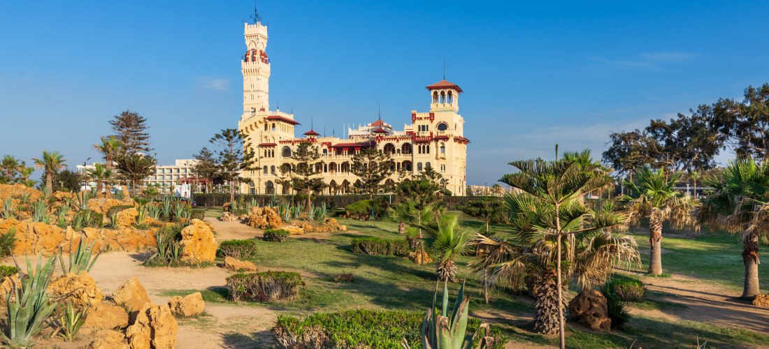 Das Bild zeigt den prächtigen Montaza Palast in Alexandria, Ägypten. Das Schloss im neoklassizistisch-orientalischen Stil mit hohen Türmen, roten Dächern und verzierten Fassaden steht im Hintergrund. Davor erstreckt sich eine gepflegte Parklandschaft mit 