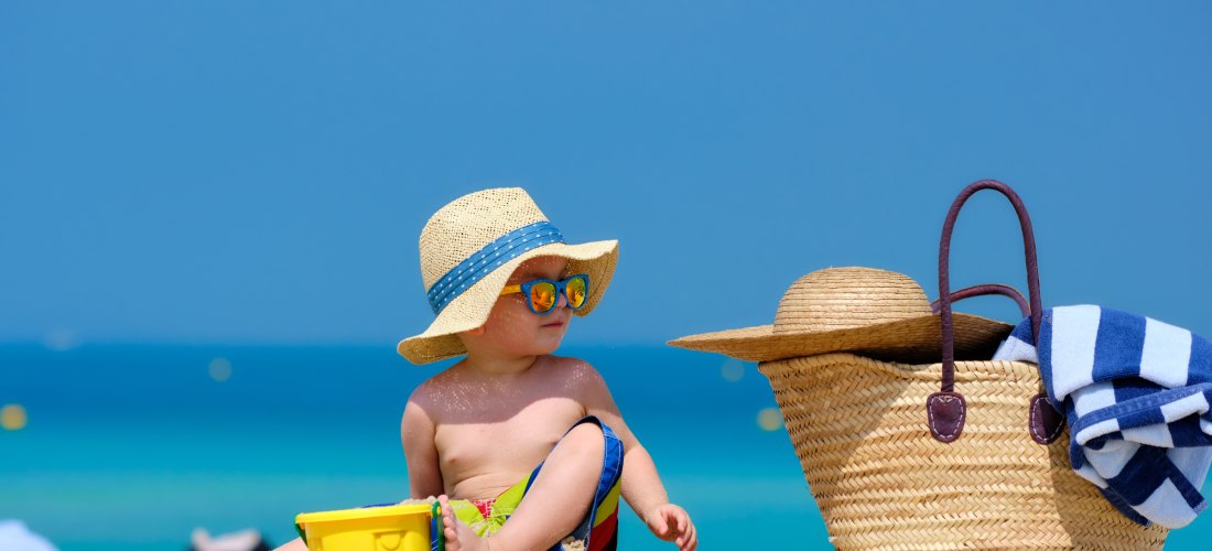 Kleinkind mit Strohhut spielt mit bunten Sandspielzeugen am Strand neben einer Strandtasche