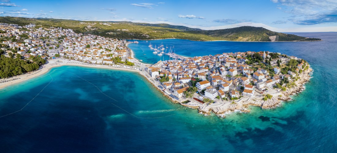 Panoramablick auf eine Küstenstadt mit dicht gedrängten Häusern mit roten Dächern auf einer Halbinsel. Rechts und links davon strahlen türkisblaues Meer und mehrere Yachten im Hafen. Im Hintergrund bewaldete Hügel.