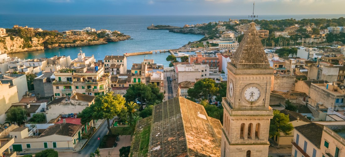 Luftaufnahme von Porto Cristo auf Mallorca. Im Vordergrund die historische Kirche Església Mare de Déu del Carme mit ihrem markanten Glockenturm und der großen Uhr. Die Kirche ist aus hellem Sandstein erbaut und steht inmitten der Altstadt mit vielen Häus