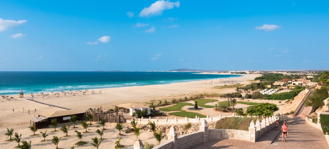 Panoramablick auf einen langen Sandstrand mit türkisfarbenem Meer auf der Insel Boa Vista, Kapverden. Sonnenliegen und Strohdächer sind über den Strand verteilt, im Vordergrund führen breite Stufen hinunter, zwei Personen gehen Richtung Wasser.