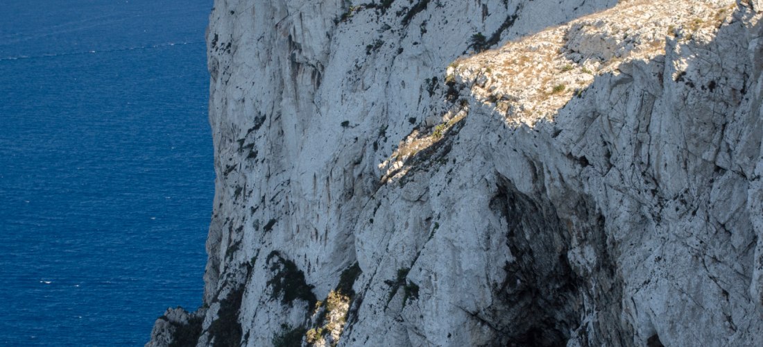 Ein weißer Leuchtturm steht auf einer imposanten, steil abfallenden Kalkstein-Klippe über dem tiefblauen Mittelmeer. Daneben befinden sich Funkmasten und einige Bäume. Die Klippe wirkt gewaltig und zerklüftet, während das Meer ruhig und grenzenlos in den 