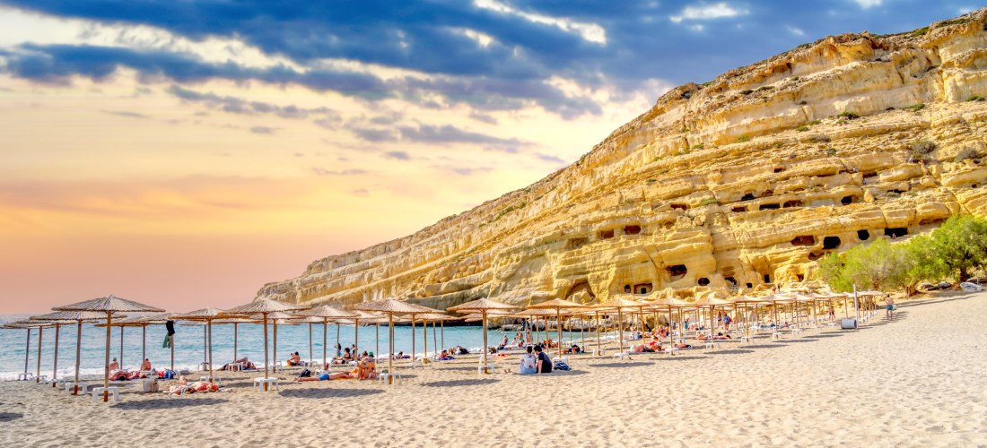 Ein breiter Sandstrand mit Liegen und Strohdächern liegt vor einer eindrucksvollen Felswand mit Höhlenöffnungen. ( Matala ) Menschen entspannen unter den Schirmen, während am Himmel ein farbenfroher Sonnenuntergang mit dunklen Wolken zu sehen ist.