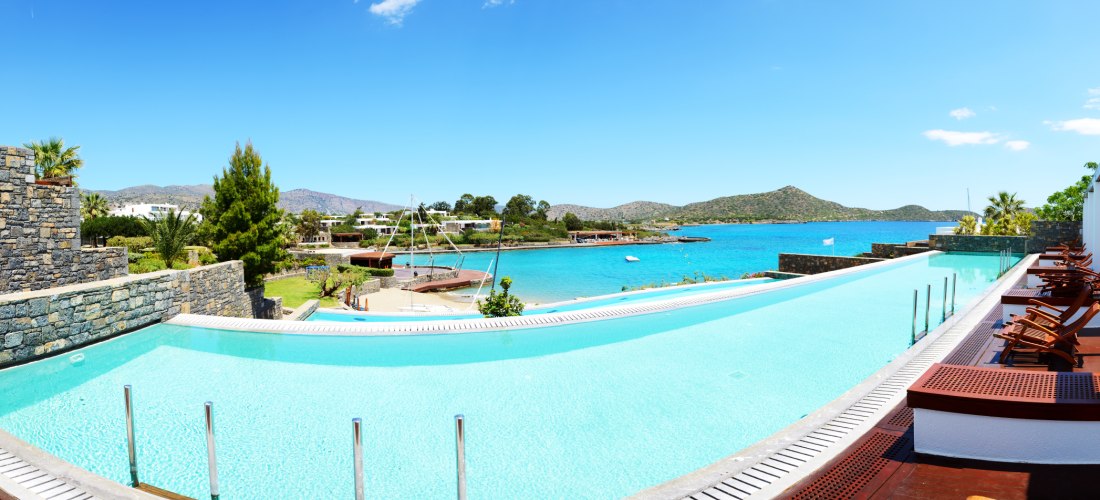 Luxus mit Meerblick: Ein moderner Infinity-Pool erstreckt sich entlang eines eleganten Resorts. Holzdielen, Liegestühle und weiße Fassaden rahmen die Szenerie. Im Hintergrund glitzert die Ägäis, eingerahmt von grünen Hügeln – ein Ort der Ruhe und Eleganz.