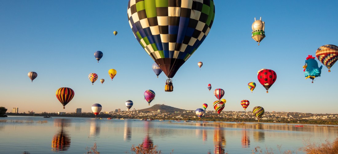 Mehrere bunte Heißluftballons steigen am frühen Morgen über einem See mit ruhigem Wasser auf, während die Stadt und Hügel im Hintergrund sichtbar sind