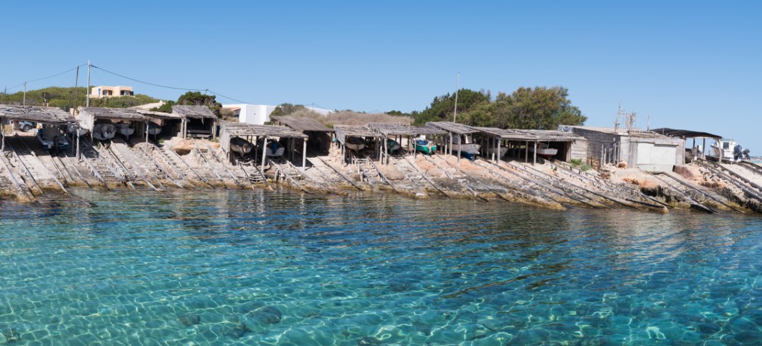 Es Caló de Sant Agustí auf Formentera: Reihe alter Fischerboothäuser mit hölzernen Sliprampen an einem kleinen Naturhafen mit kristallklarem, türkisfarbenem Wasser; links ein weißes Haus, sonniger Himmel.