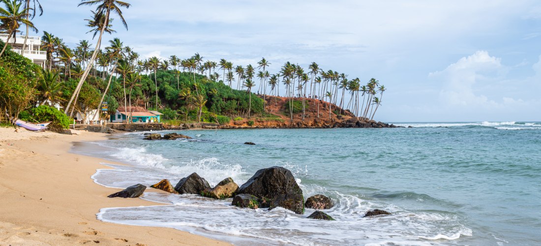 Tropischer Sandstrand mit Palmen und Felsen in Mirissa, Sri Lanka, mit sanften Wellen des Indischen Ozeans