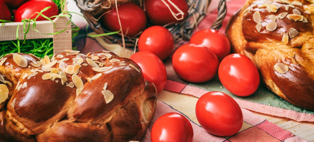 Nahaufnahme eines traditionellen griechischen Ostergebäcks, mit goldbrauner, glänzender Kruste und Mandelblättchen bestreut. Rundherum liegen zahlreiche hartgekochte, kräftig rot gefärbte Eier, teilweise in Körbchen mit Stroh oder auf einer karierten Tisc
