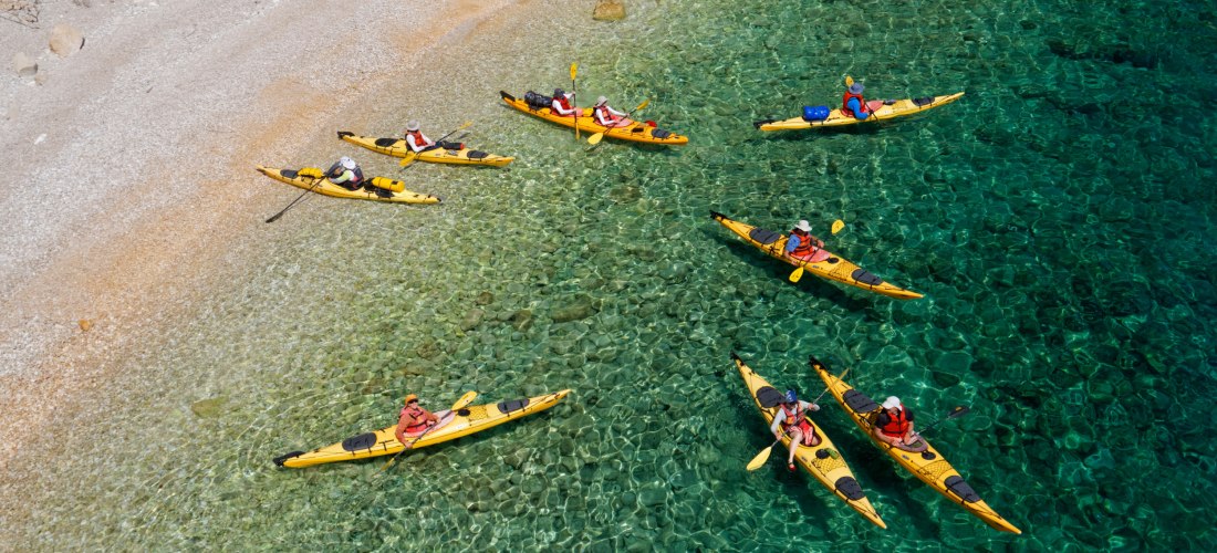 Gruppe von Menschen in gelben Kajaks auf kristallklarem, türkisfarbenem Wasser in der Nähe eines steinigen Strandes. Die Paddler tragen Schwimmwesten und Sonnenhüte.