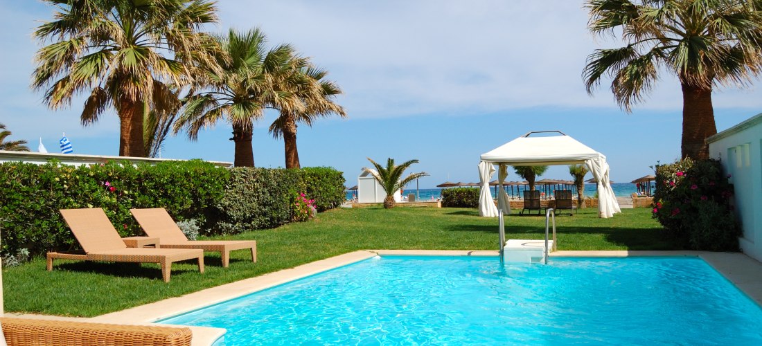 Ein privater Gartenbereich mit zwei Sonnenliegen, einem kleinen Pool und einem weißen Pavillon mit Blick auf das Meer. Palmen, gepflegter Rasen und bunte Blumen umgeben den Pool. Im Hintergrund sieht man den Strand mit Sonnenschirmen und das türkisfarbene