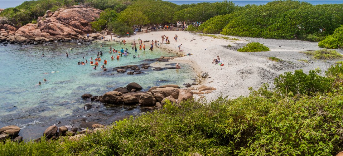 Badegäste am Strand und im klaren Wasser von Pigeon Island bei Trincomalee, Sri Lanka, umgeben von Felsen und Mangroven