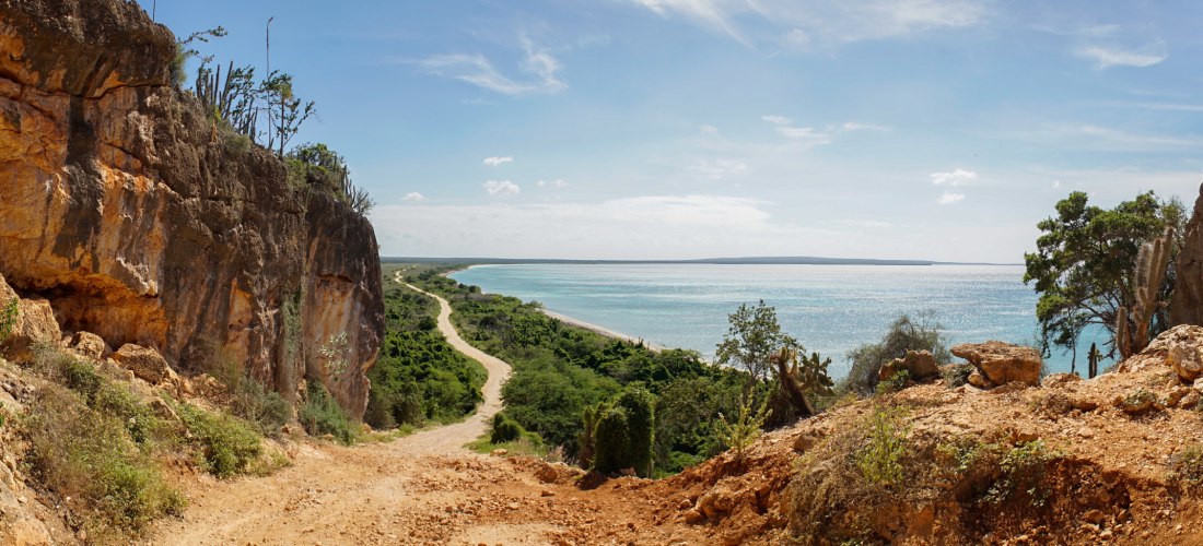 Panoramablick von einem felsigen Küstenpfad auf eine tropische Landschaft mit grüner Vegetation, Sandweg und türkisfarbenem Meer