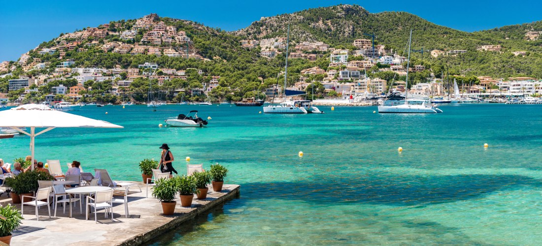 Sonnige Küstenlandschaft mit kristallklarem türkisfarbenem Wasser. Im Vordergrund eine Terrasse mit weißen Tischen und Stühlen, auf der Menschen unter weißen Sonnenschirmen sitzen. Im Hintergrund grüne Hügel mit vielen Häusern, sowie mehrere Segelboote un