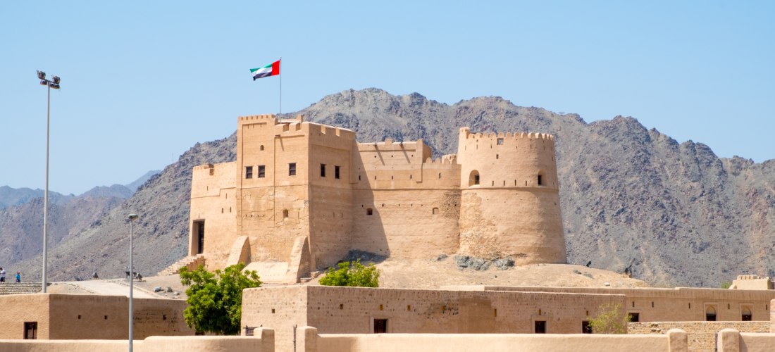 Außenansicht des Fujairah Fort in den Vereinigten Arabischen Emiraten. Historische Festung mit Wachtürmen und Flagge, vor der Kulisse des Hadschar-Gebirges.