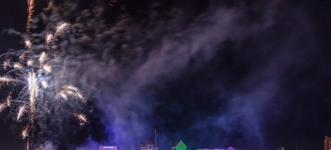 Nächtliches Feuerwerk über der bunt beleuchteten Häuserzeile von Willemstad, Curaçao. Links steigen silberne Raketen in den Himmel, während sich am Wasser bunte Lichter spiegeln. Dichte Rauchschwaden ziehen über die Szenerie.