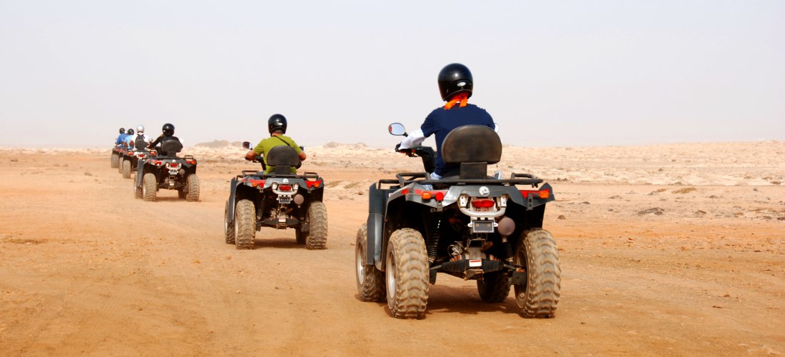 Gruppe von Menschen auf Quads fährt in einer Reihe durch eine weite, staubige Wüstenlandschaft. Die Fahrenden tragen Helme, die Umgebung ist flach und trocken, der Himmel klar.
