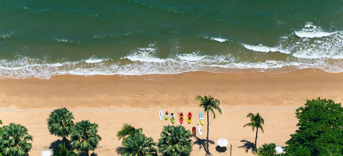 Luftaufnahme eines tropischen Strandes mit Palmen, bunten Kajaks und Stand-up-Paddle-Boards im Sand. Kleine Wellen treffen auf den goldenen Sand, das Wasser schimmert grünlich-blau
