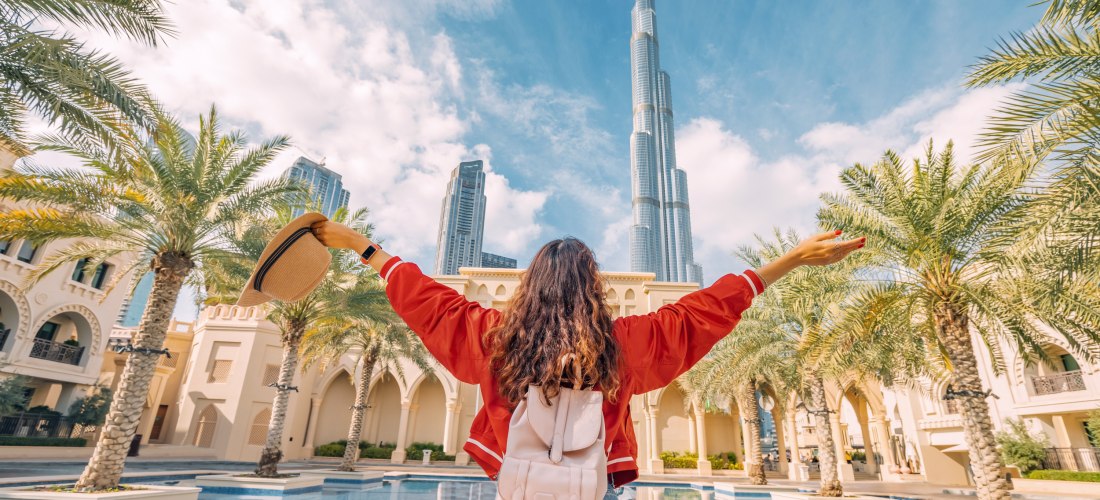 Eine junge Frau mit Rucksack steht vor einem Palmenhof und blickt mit ausgestreckten Armen auf den Burj Khalifa in Dubai, den höchsten Wolkenkratzer der Welt. Sie trägt eine rote Jacke und hält einen Sonnenhut in der Hand. Der Himmel ist blau mit einigen 