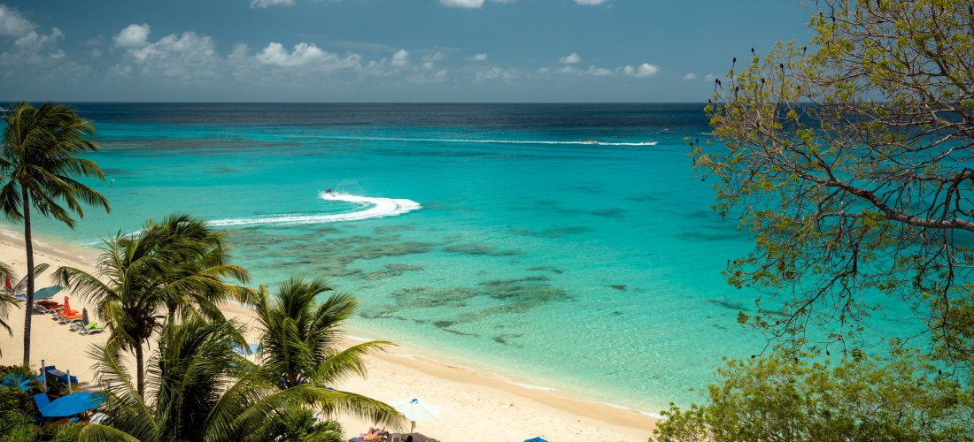 Blick auf einen weißen Sandstrand auf Barbados mit türkisfarbenem Meer. Palmen säumen den Strand, im Wasser zieht ein Jetski eine weiße Spur. Einige Sonnenliegen mit blauen Schirmen stehen im Schatten der Bäume, der Himmel ist teils bewölkt.