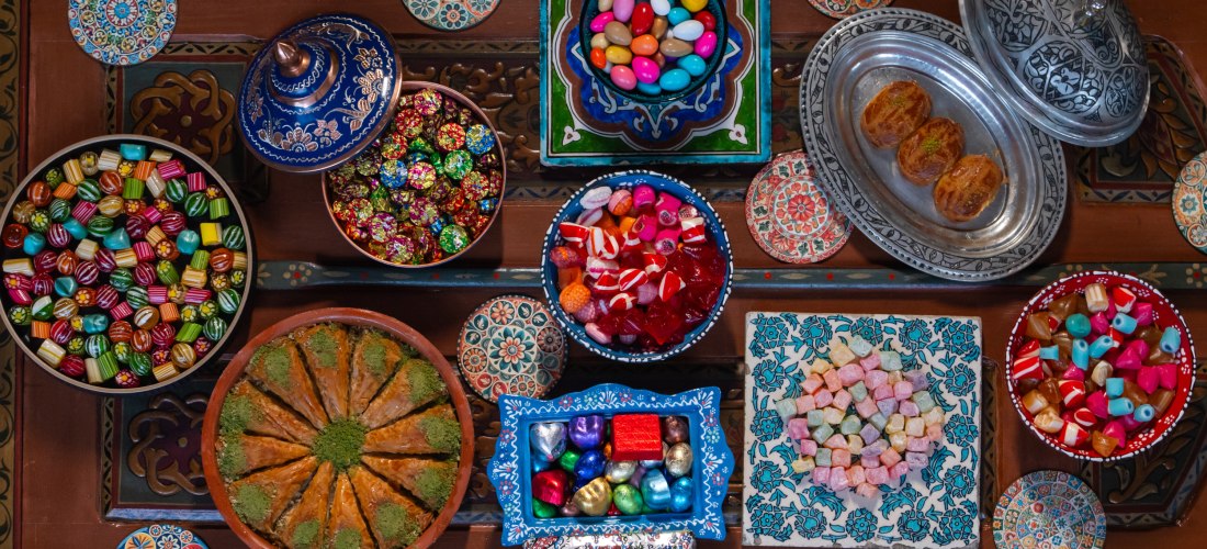 Verschiedene bunte Süßigkeiten und traditionelle Desserts sind kunstvoll in Schalen auf einem orientalisch verzierten Tisch angerichtet. Darunter Baklava, gefüllte Bonbons, Lokum und Pralinen – alles in festlicher Atmosphäre.