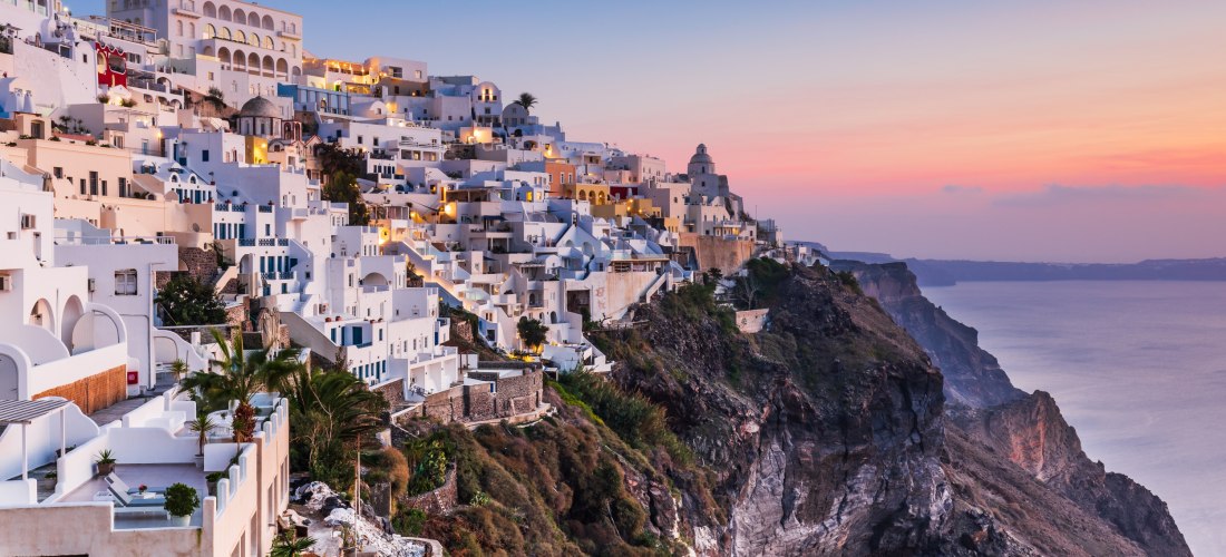 Fira bei Sonnenuntergang: weiß getünchte Häuser kleben an der Kraterkante, rosa-blauer Himmel über der Ägäis