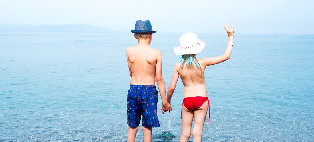 Zwei Kinder stehen Hand in Hand im seichten Wasser am Kiesstrand und blicken aufs Meer hinaus. Das Kind links trägt eine blaue Badehose und einen blauen Hut, das Kind rechts einen weißen Sonnenhut und einen roten Bikini. Es hebt die freie Hand und zeigt e