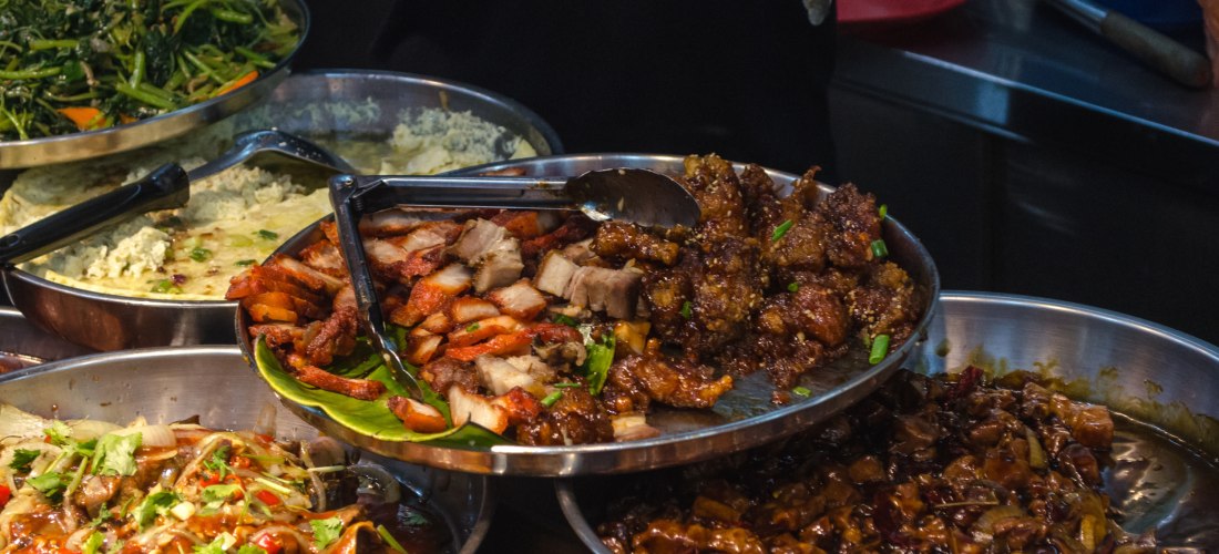 Nahaufnahme einer Auswahl an Streetfood in Singapur. In mehreren Edelstahlschalen befinden sich gebratene Fleischstücke, würzige Soßen, gebratenes Gemüse und aromatische asiatische Gerichte. Ein Koch im Hintergrund bedient das Buffet mit einer Zange