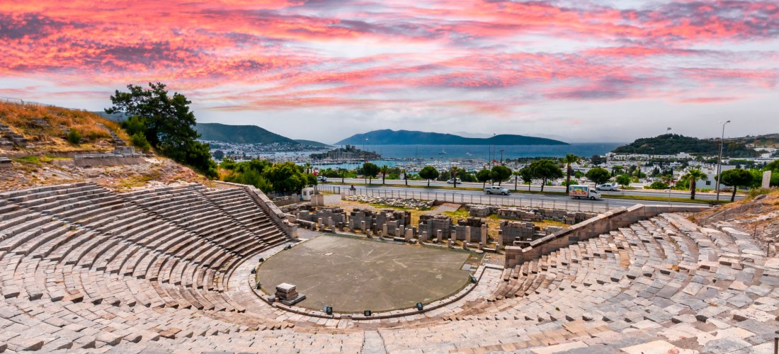 Antikes Amphitheater in Bodrum, Türkei, mit halbkreisförmigen Steinstufen und Blick auf die Stadt, das Meer und bewaldete Hügel. Der Himmel leuchtet in dramatischem Rosa und Orange bei Sonnenuntergang.
