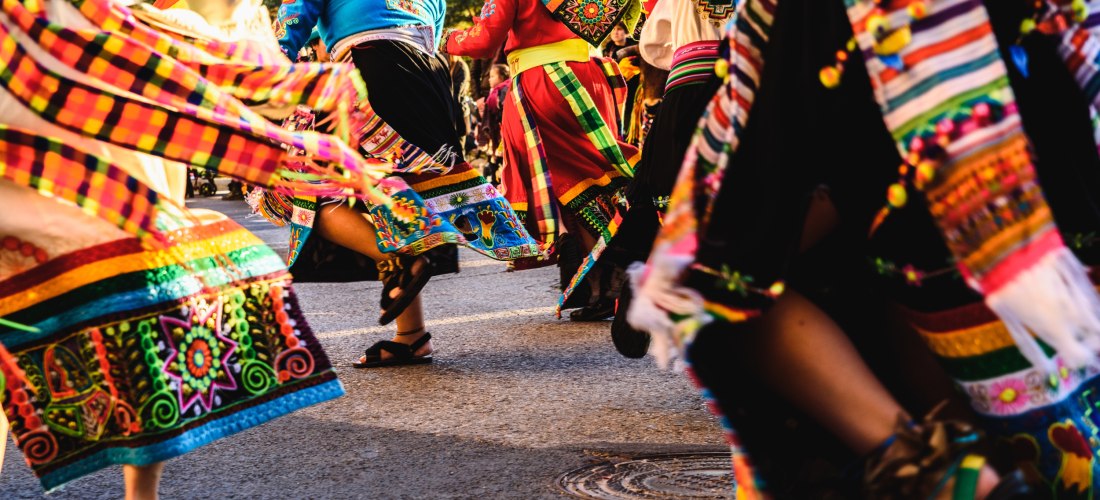 Eine Gruppe von Tänzerinnen und Tänzern in bunten, traditionellen Gewändern tanzt auf einer Straße. Die Kleidung ist mit Stickereien, Bändern und farbenfrohen Mustern verziert. Die Szene ist dynamisch und lebendig, die Tänzer sind in Bewegung, die Röcke u