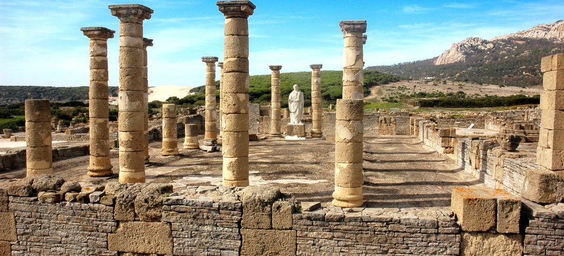 Römische Ruinen von Baelo Claudia nahe der Playa de Bolonia. Mehrere erhaltene Steinsäulen stehen in Reih und Glied, im Hintergrund eine weiße Statue, dahinter Dünen und grüne Hügel unter blauem Himmel.