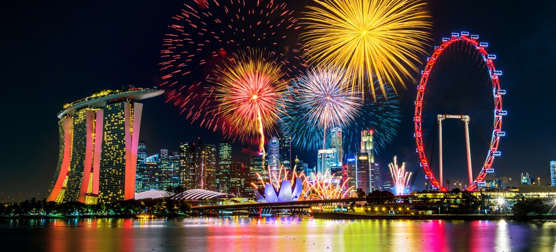 Farbenfrohes Feuerwerk über der Marina Bay in Singapur. Zu sehen sind das markante Hotel Marina Bay Sands, das rot beleuchtete Riesenrad Singapore Flyer und zahlreiche Hochhäuser der Skyline. Die Spiegelung der Lichter im Wasser rundet die nächtliche Szen