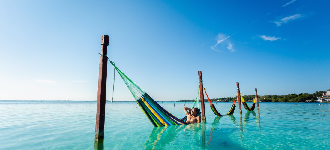 Mann entspannt in einer bunten Hängematte über türkisblauem Wasser, befestigt an Holzpfosten, unter einem klaren blauen Himmel