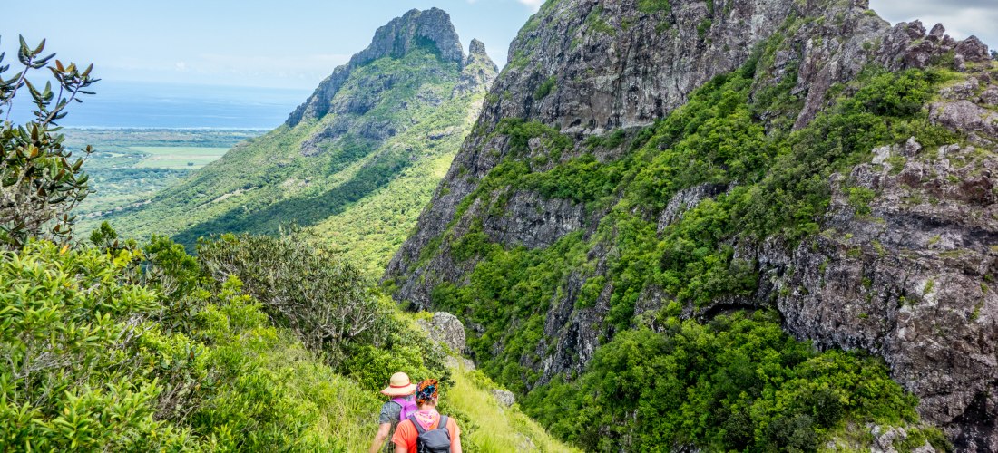 Dieses Bild zeigt eine abenteuerliche Wanderung durch eine atemberaubende Berglandschaft. Zwei Wanderer, ausgestattet mit Rucksäcken und Hüten, bewegen sich vorsichtig auf einem schmalen, grasbewachsenen Pfad entlang eines steilen Hangs. Die Berge sind ei