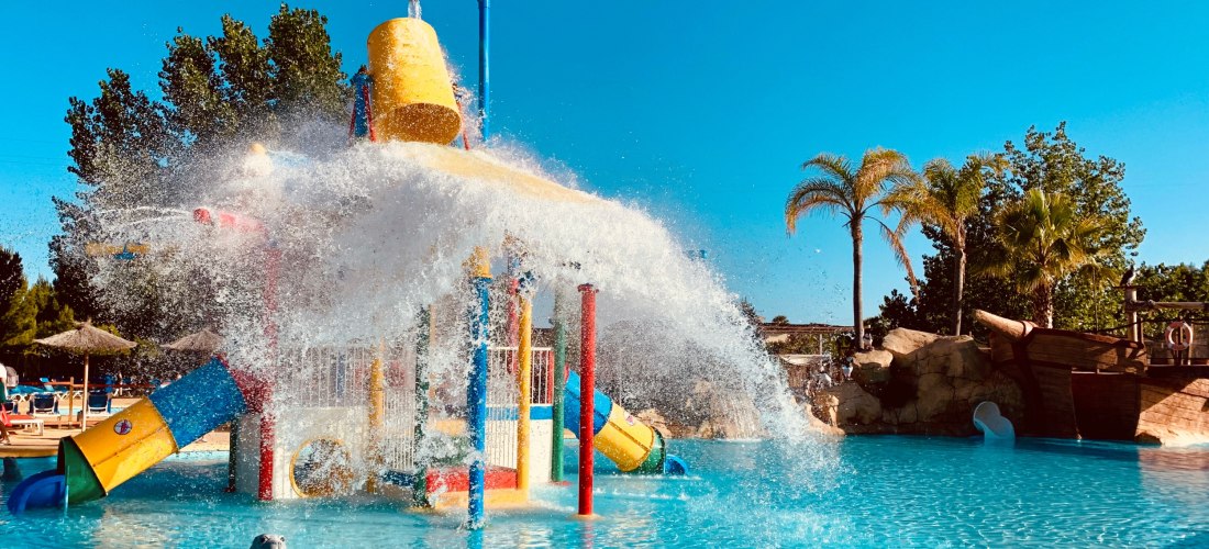 Ein großer Kippeimer schüttet eine Wasserflut über ein buntes Kletter- und Spielgerüst in einem flachen Becken; dahinter Palmen, Sonnenschirme und ein Bereich im Piratenschiff-Stil unter blauem Himmel.