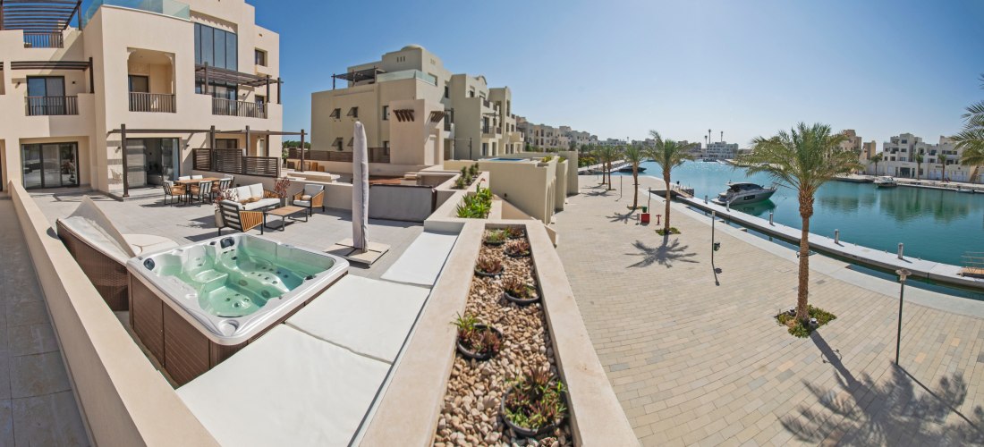 Moderne Apartments mit Terrasse und Whirlpool an einer Marina mit Palmenpromenade und Yachten im Wasser. Klare blaue Himmel, ruhige Urlaubsatmosphäre