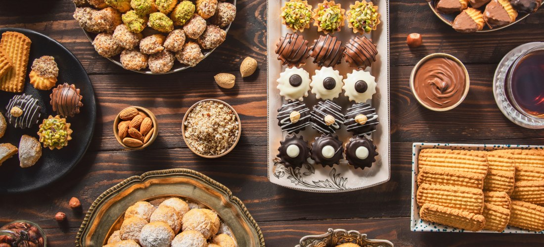 Eine reich gedeckte Holztafel mit verschiedenen orientalischen Süßigkeiten wie gefüllten Keksen, Gebäck mit Pistazien, Schokoladentrüffeln und Mandeln. Dazwischen kleine Schälchen mit Nüssen, Datteln und Schokoladencreme