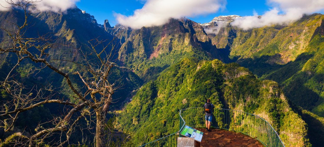 Aussichtsplattform über einer üppig grünen Berglandschaft (Madeira): Eine Person blickt auf zerklüftete, teils wolkenverhangene Gipfel unter blauem Himmel.
