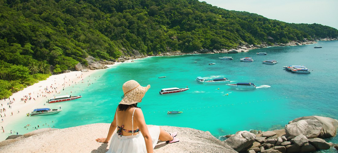 Eine Frau mit Sonnenhut, Bikini und weißem Rock sitzt auf einem Felsen oberhalb einer tropischen Bucht mit türkisfarbenem Wasser. Sie blickt auf einen weißen Sandstrand, an dem viele Menschen baden und entspannen. Im Wasser ankern mehrere Schnellboote und