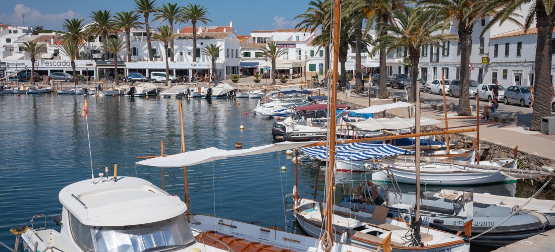 Hafenpromenade im Ort Fornells auf Menorca mit weißen Häusern, Palmen und zahlreichen Fischerbooten, die am Kai vertäut sind.