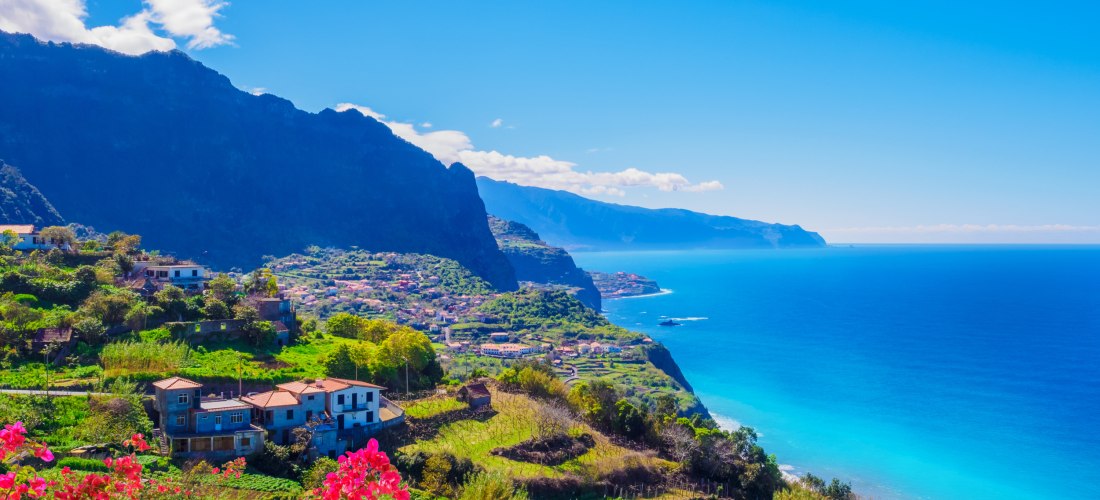 Panoramablick auf die grüne Nordküste Madeiras mit blühenden roten Bougainvillea im Vordergrund. Dahinter liegen kleine Dörfer auf terrassierten Hängen, steile Klippen und das tiefblaue Meer, das sich bis zum Horizont erstreckt.