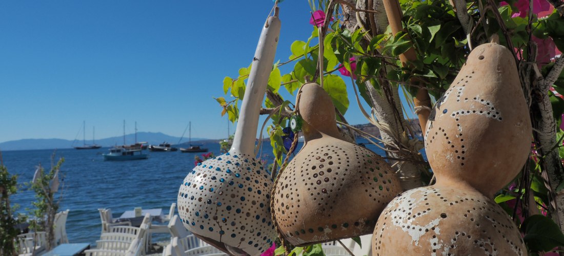 Handverzierte Trockenfrüchte-Lampen hängen an einer blühenden Pflanze vor einem kleinen Restaurant direkt am Meer. Im Hintergrund sieht man weiße Stühle, Tische und segelnde Boote auf dem ruhigen Wasser unter klarem, blauem Himmel.