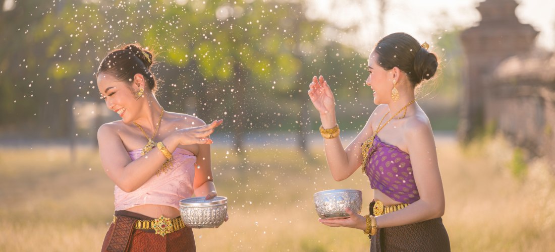 Zwei Frauen in traditioneller thailändischer Kleidung spielen lachend mit Wasser im Sonnenlicht. Beide halten verzierte Silberschalen und tragen goldenen Schmuck. Um sie herum fliegen glitzernde Wassertropfen.