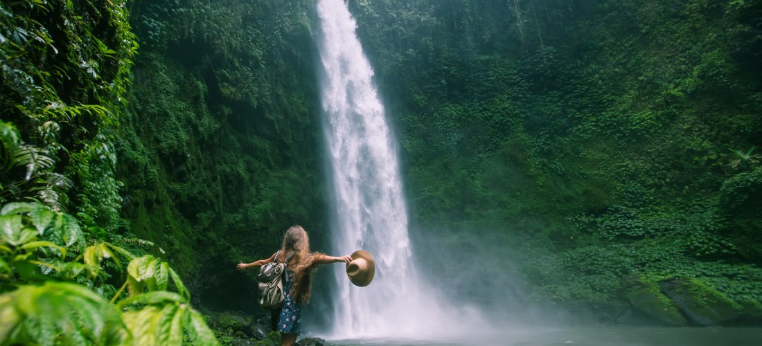 Eine Frau mit langem Haar, Sommerkleid und Rucksack steht barfuß auf nassen Steinen vor einem hohen Wasserfall inmitten tropischer Vegetation auf Bali. Sie hält ihren Hut in der Hand und breitet die Arme aus, während Wassernebel die Luft durchzieht. Der W