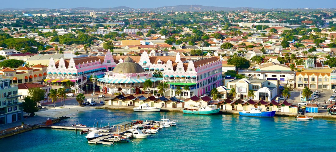 Luftaufnahme der Altstadt von Oranjestad auf Aruba. Im Vordergrund liegt der Hafen mit Booten, dahinter bunt verzierte Gebäude mit Giebeldächern und einem auffälligen runden Kuppelbau. Die Stadt erstreckt sich bis zu den Hügeln am Horizont.