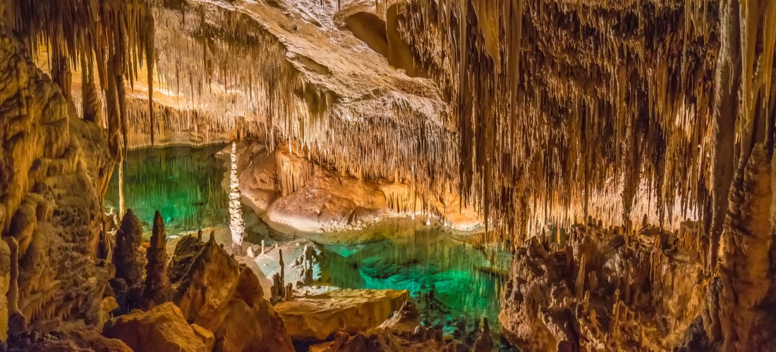Beleuchtete Tropfsteinhöhle mit Stalaktiten und Stalagmiten sowie einem türkisfarbenen unterirdischen See in den Drachenhöhlen auf Mallorca