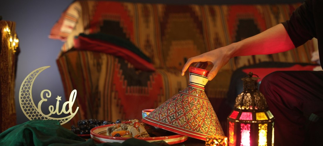 Ein Mensch hebt den Deckel einer kunstvoll bemalten Tajine bei festlicher Beleuchtung. Daneben orientalische Laternen, eine goldene 'Eid'-Dekoration in Halbmondform und ein Teller mit traditionellem Essen. Im Hintergrund ein gemusterter Sofaüberwurf.
